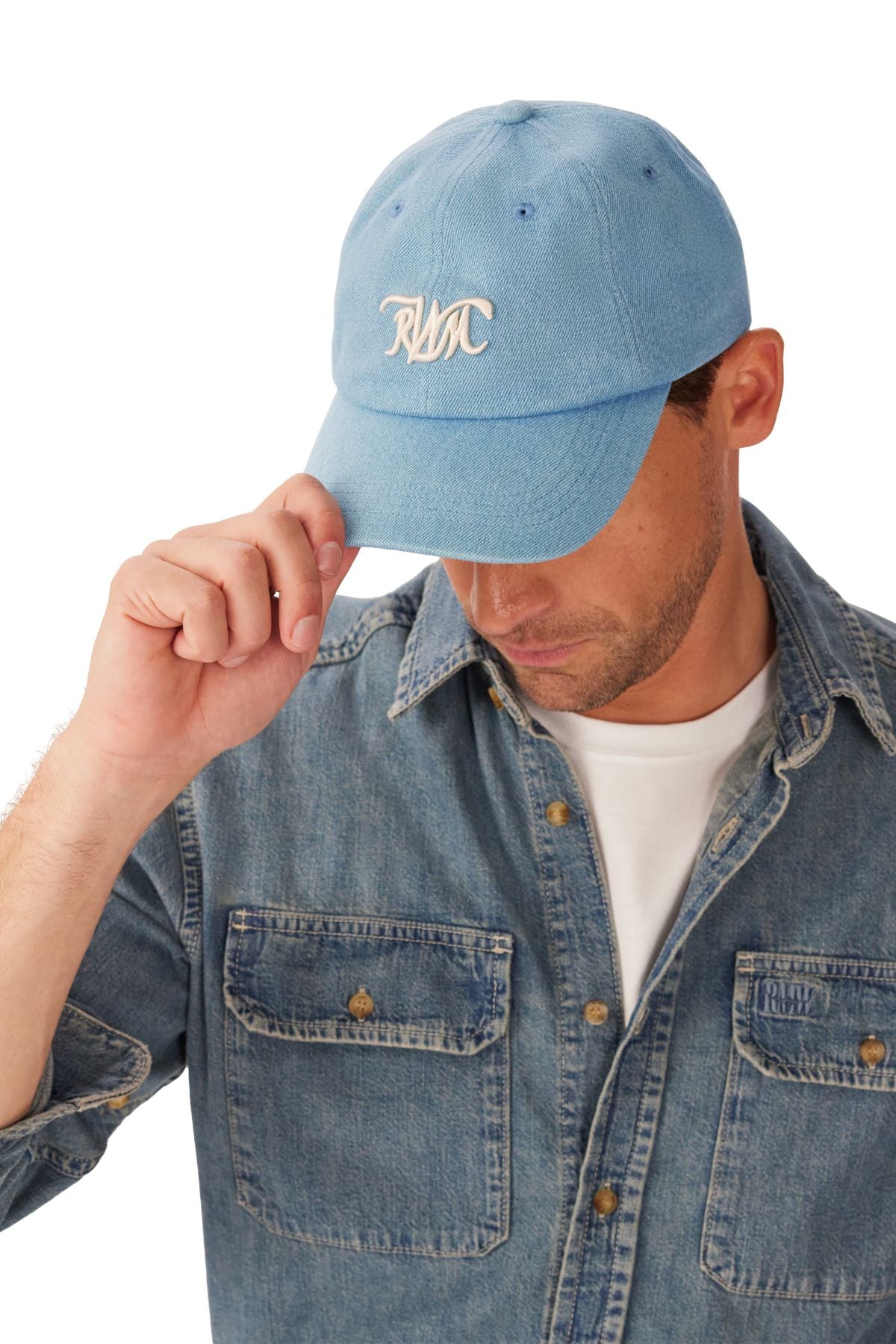 A man wearing an RM Williams Cursive Twill Cap and a denim button-up shirt over a white t-shirt looks down, adjusting the cap's brim with one hand.