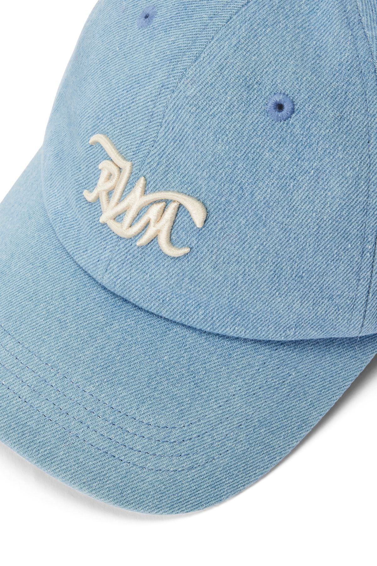 The RM Williams Cursive Twill Cap is a light blue organic cotton cap with a curved brim, white embroidered "RMC" text on the front, visible stitching, and two crown ventilation eyelets.