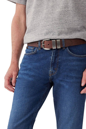 A person wearing a gray t-shirt tucked into blue jeans, secured with the RM Williams Drover Belt, which is handmade in Australia from brown leather and features a silver buckle along with additional metal detailing. The image focuses on the outfit from the waist to mid-thigh.