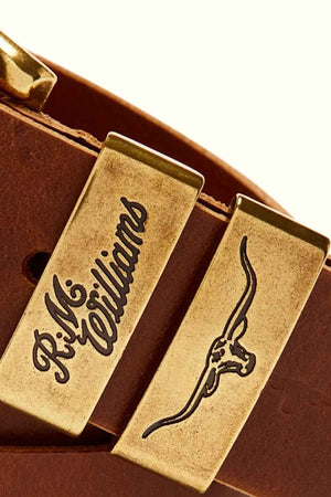 Close-up of the RM Williams Drover Belt, a brown pull-up leather belt featuring two gold-toned metal accents. One accent is engraved with the text "R.M. Williams," and the other features an embossed longhorn bull design. Made in Australia, this belt showcases fine craftsmanship and attention to detail.