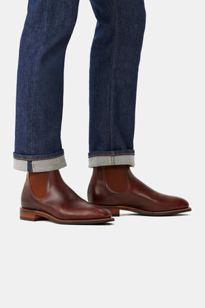 A person wears RM Williams Dynamic Flex Craftsman handcrafted leather Chelsea boots with a leather and rubber sole, paired with dark blue jeans with rolled cuffs, standing out against a plain light gray background.