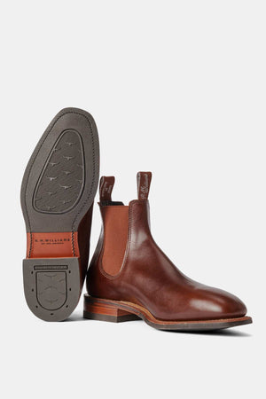 The RM Williams Dynamic Flex Craftsman Boot features handcrafted leather, elastic side panels, and pull tabs. One boot stands upright, while the other displays a leather and rubber sole with brand logos and patterns. It has a sleek design complemented by a wooden heel.