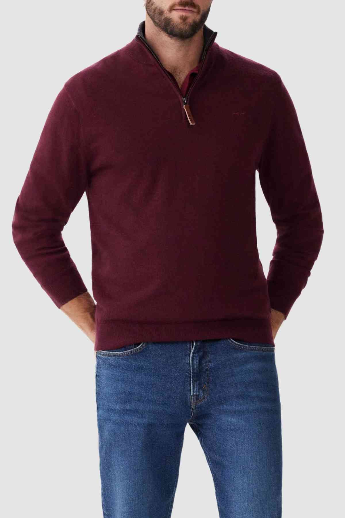 A person wearing the RM Williams Ernest Sweater, a maroon fine gauge merino quarter-zip featuring a dark zipper with a brown leather pull tab, and blue jeans stands with hands in pockets against a plain, light gray background. The person’s face is not visible.