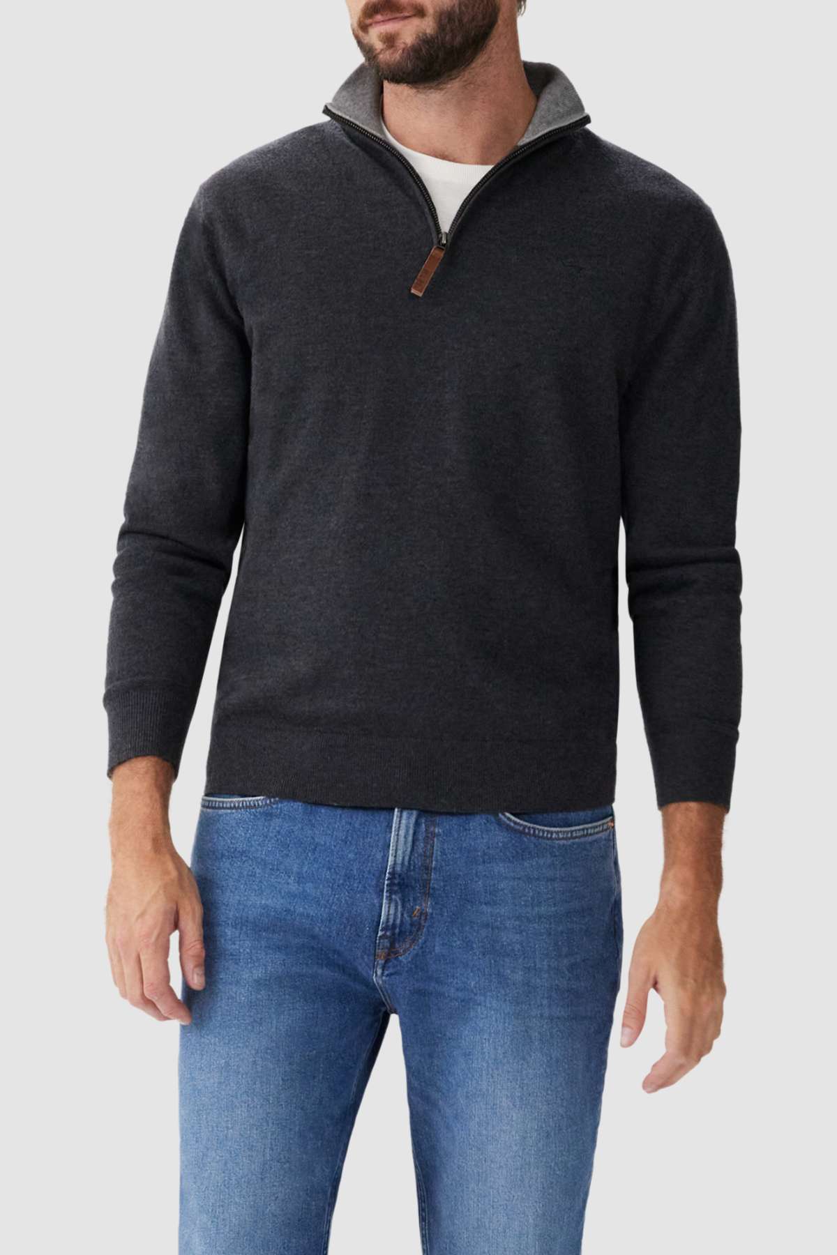 A person is wearing the RM Williams Ernest Sweater in dark gray over a white t-shirt and blue jeans. The fine gauge merino quarter-zip sweater features a small brown zipper pull and a high collar, styled against a plain light gray background.