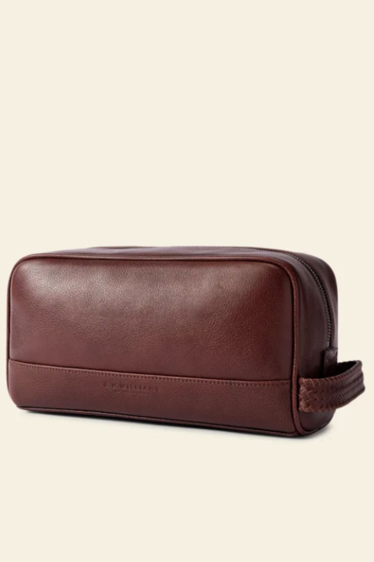 The RM Williams Farrier Signature Washbag is a brown tumbled grain leather travel toiletry bag with a rectangular shape, subtle stitching, top zipper, and woven end handle, shown against a plain beige background.