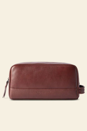 The RM Williams Farrier Signature Washbag is a rectangular brown tumbled grain leather toiletry bag with a zipper, side handle, and water-resistant lining, shown against a plain beige background.