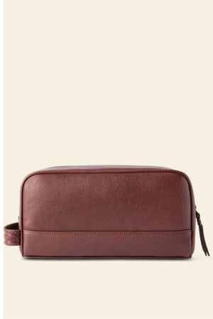 The RM Williams Farrier Signature Washbag is a rectangular brown tumbled grain leather toiletry bag with a zipper closure, water-resistant lining, and side handle, shown on a beige background.