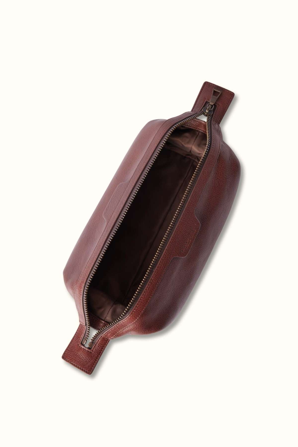 The RM Williams Farrier Washbag is crafted from tumbled grain leather, featuring a textured brown surface. It has a zippered opening that reveals a spacious interior and includes a side handle for easy carrying.