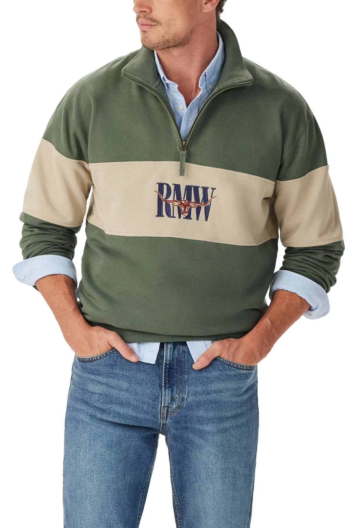 A man wearing a green and beige RM Williams Heritage 1/4 Zip Sweatshirt with "PMV RW" on the front, a light blue collared shirt underneath, and blue jeans has his hands in his pockets.