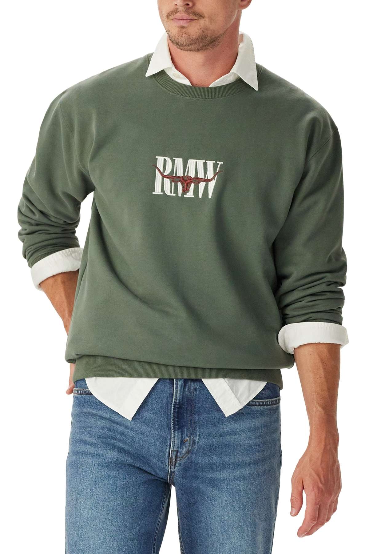 A man wears the RM Williams Heritage Crew Sweatshirt in green with "RM W" in white on the front, styled over a white collared shirt and blue jeans. He has his sleeves rolled up and stands with one hand in his pocket.