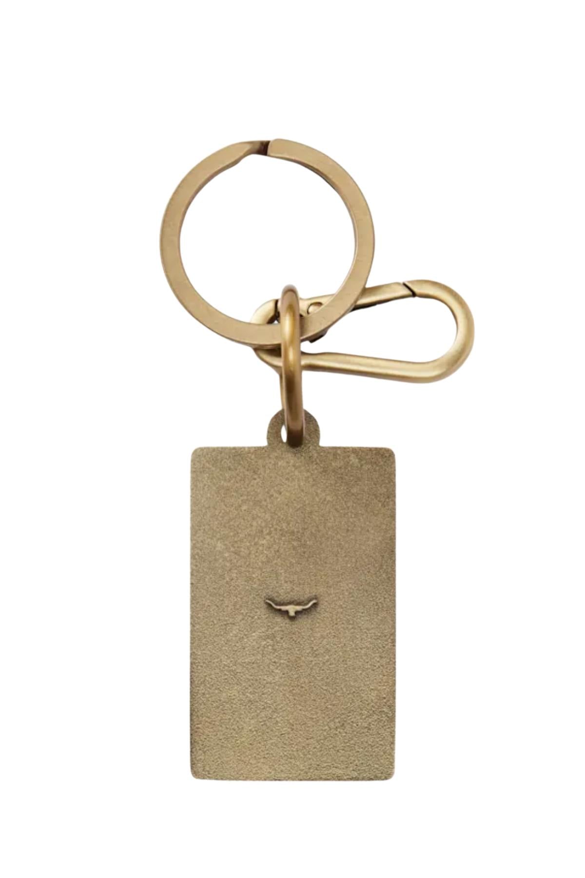 The RM Williams Heritage Logo Enamel Key Ring features a rectangular brass keychain with an engraved bird logo, attached to a round enamel key ring and clasp, with brass hardware contrasting against the white background.