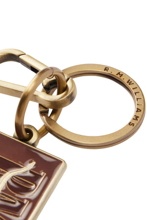 Close-up of the RM Williams Heritage Logo Enamel Key Ring, engraved with "R. M. WILLIAMS," attached to a carabiner and a brown rectangular tag featuring gold accents and sturdy brass hardware.