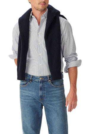 A man wearing the RM Williams Ingham Poplin Stripe Shirt with rolled-up sleeves, blue jeans, and a dark ribbed sweater draped over his shoulders, with the sweater’s sleeves loosely tied in front.