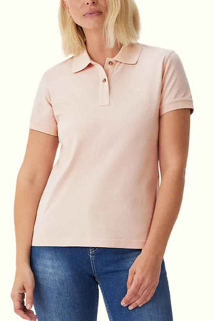 A woman in a light pink RM Williams Margaret Polo shirt, made from pique cotton with a ribbed collar, stands with one arm slightly bent. Her light bob-styled hair adds to the look, completed by blue jeans against a plain white background.