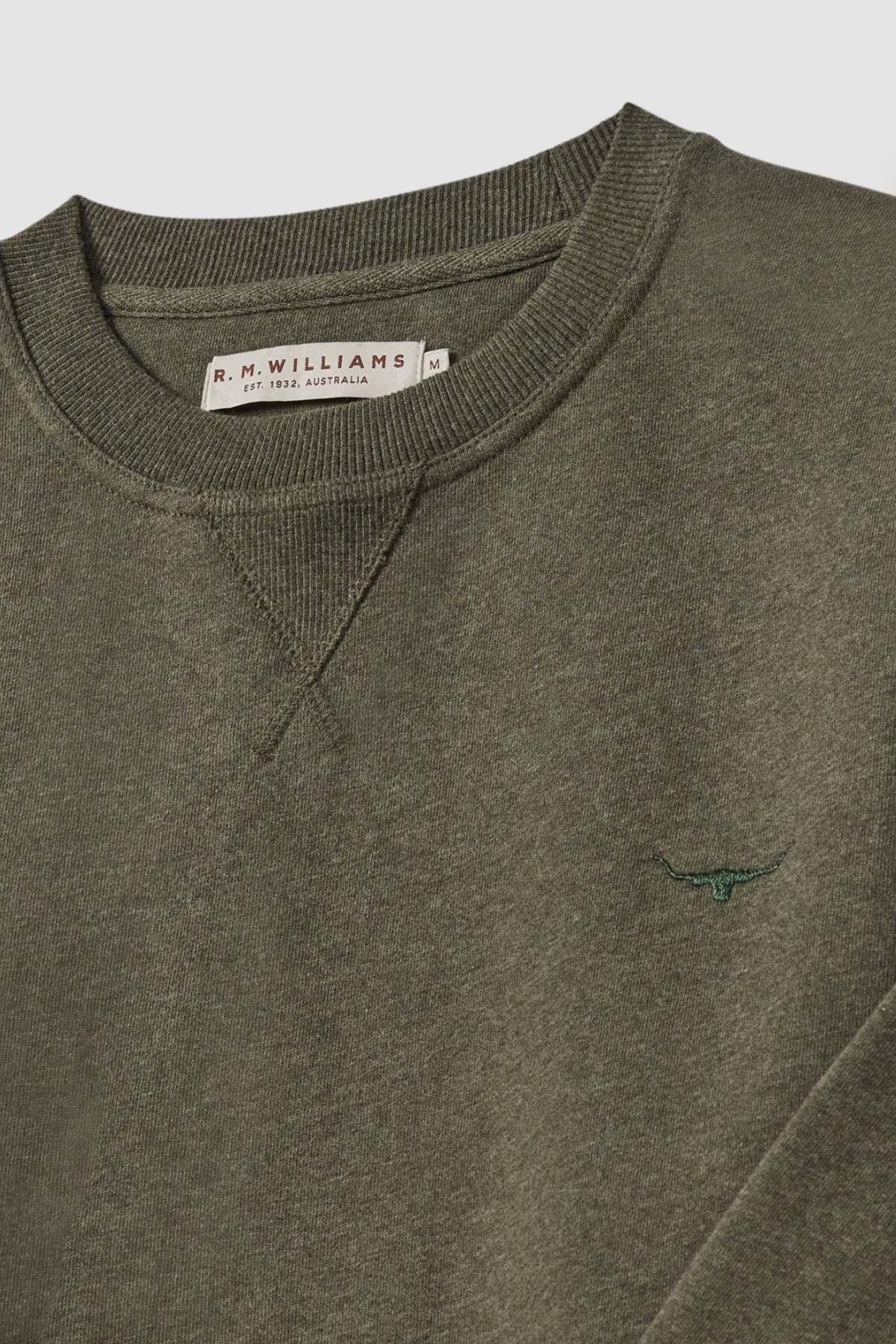 A close-up of a green RM Williams Mini Longhorn Sweatshirt. The label inside the collar reads "R.M. Williams Est. 1932 Australia S." A small embroidered longhorn logo is visible on the chest, depicting a bull's head. The recycled cotton poly fleece fabric appears to be soft and comfortable.