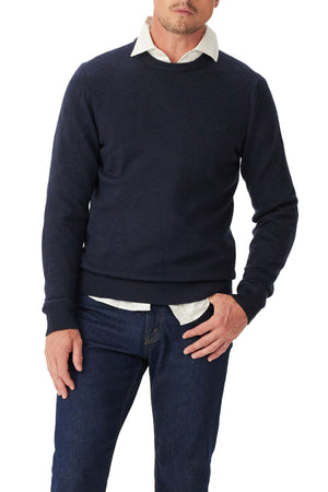 A man wearing the RM Williams Murphy Merino Crew Knit in navy over a white collared shirt and dark blue jeans stands with one hand in his pocket against a white background.