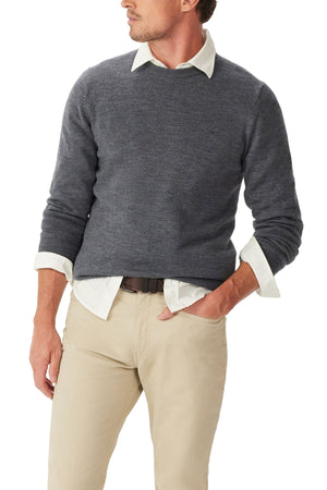 A man models the RM Williams Murphy Merino Crew Knit, a dark gray sweater layered over a light collared shirt with beige pants. One hand is in his pocket as he looks to the side. The image shows him from shoulders to mid-thigh.