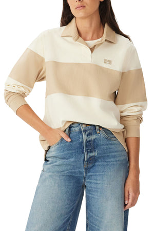 A person wearing the RM Williams Narran 4 Panel Rugby, a long-sleeve shirt with wide beige and cream stripes, a collar, and "Rugby RL67" embroidery on the chest, paired with blue jeans and one hand in their pocket.
