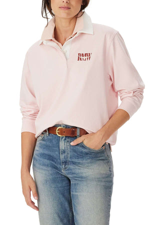 A person wearing the RM Williams Narran Heritage Rugby in light pink, with "DMV" text on the chest, blue jeans, and a brown belt. One hand is in their pocket and their face is partially out of frame.