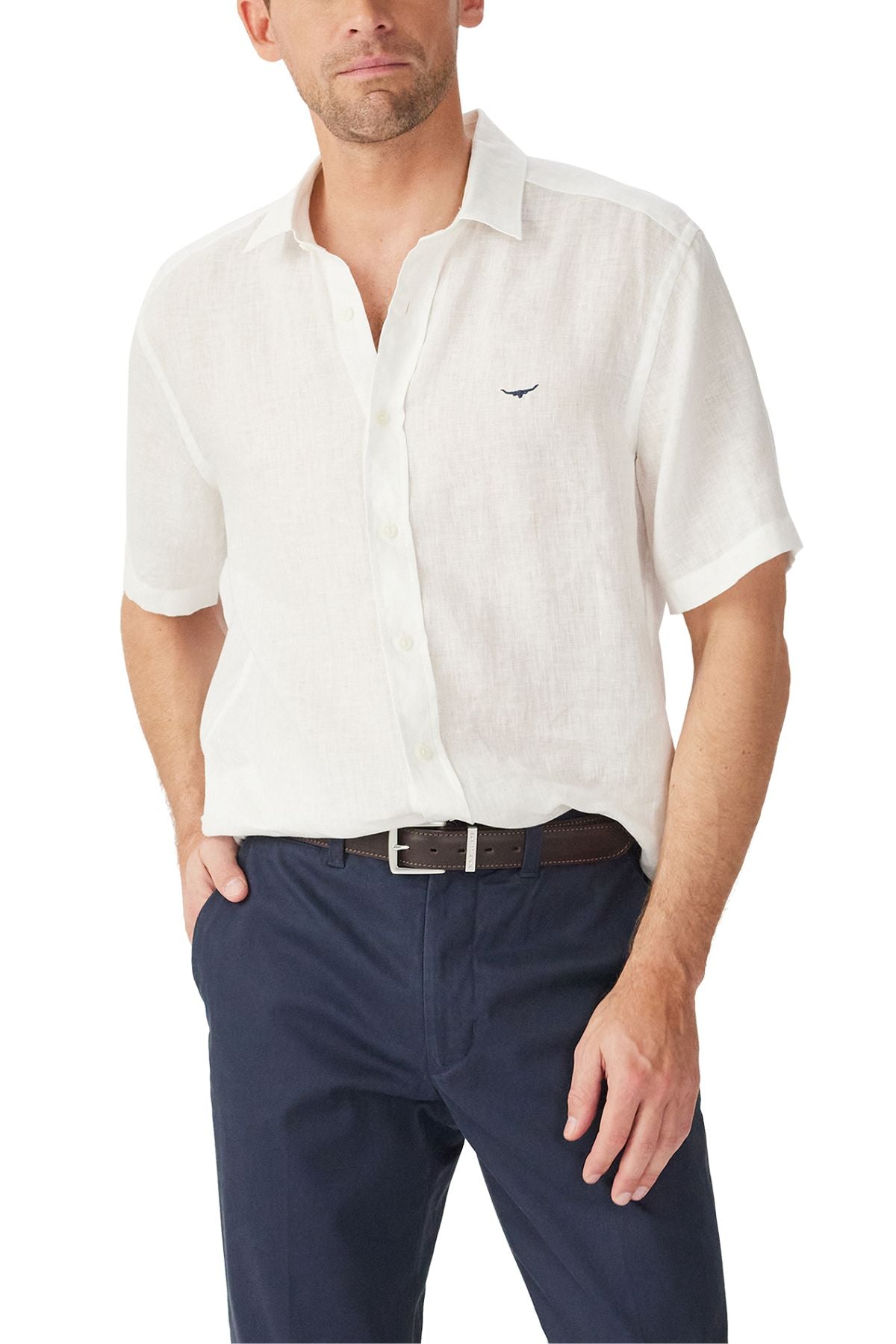 A man wears the RM Williams Newman Linen Short Sleeve Shirt in white, tucked into navy blue pants with a dark belt. The relaxed fit shirt features French linen and a small embroidered logo on the chest. His left hand rests in his pocket.