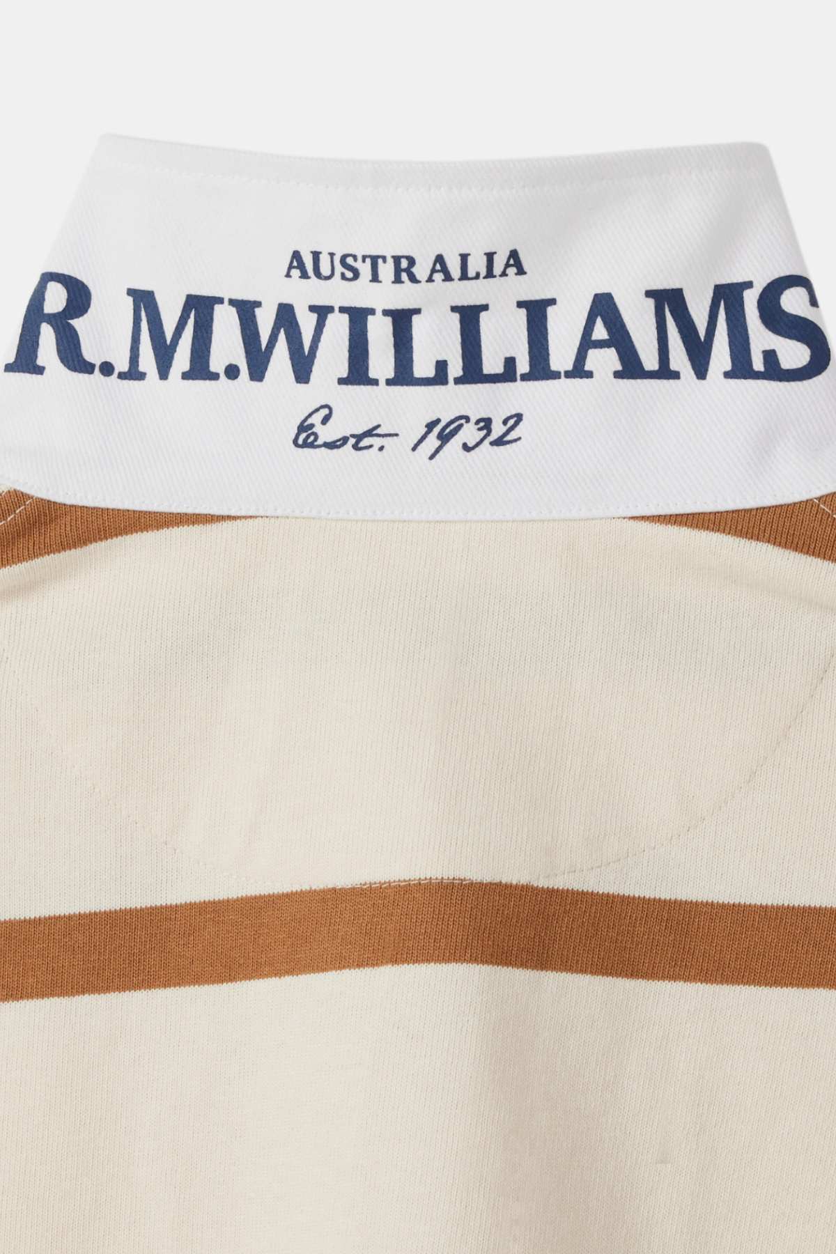 Close-up of a white collar with "R.M. Williams" in large blue text and "Australia Est. 1932" in smaller blue text on the RM Williams Nundle Rugby shirt. The shirt is beige with a thick horizontal brown stripe running across the back just below the collar.