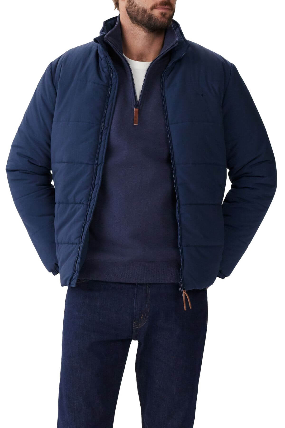 Man in RM Williams Padstow Jacket, crafted from recycled nylon with Primaloft fill, wearing a navy quarter-zip sweatshirt and dark jeans. He stands with hands in the jacket pockets against a plain white background.
