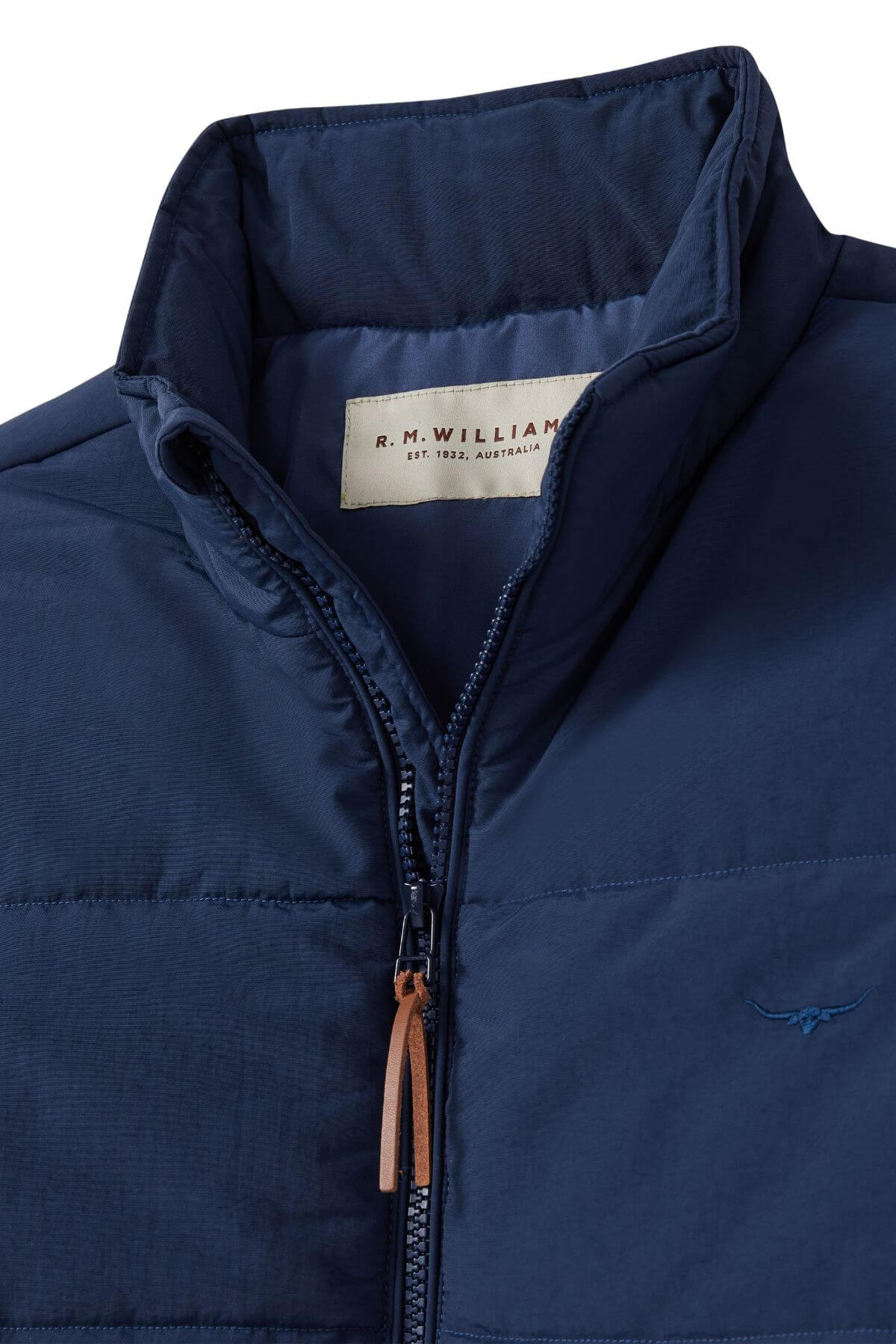 Close-up of the RM Williams Padstow Jacket, a dark blue puffer with Primaloft fill and high collar. Features a brown leather zipper pull and an embroidered longhorn logo on the chest. Made from recycled nylon with an inside label: "R.M. Williams, Est. 1932, Australia.
