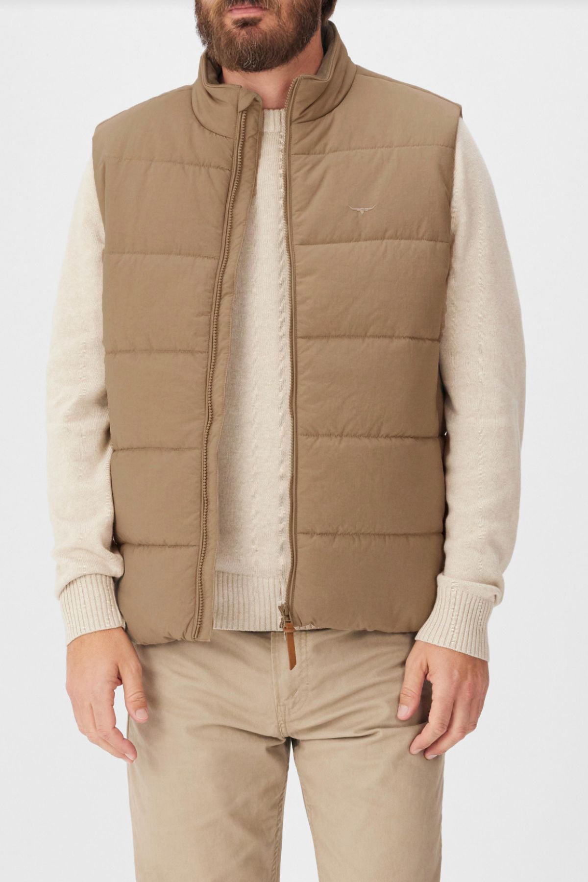 A man wears the RM Williams Padstow Vest—a beige puffer made from recycled nylon—over a light beige sweater and beige pants. He stands with arms relaxed at his sides against a plain background, head partially out of frame.