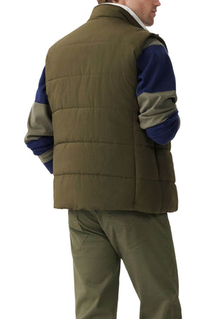 A person is standing with their back to the camera, showcasing an RM Williams Padstow Vest in olive green with PrimaLoft filling over a long-sleeved, color-blocked shirt in navy, gray, and green. Matching olive green pants complete the ensemble against a plain white background.