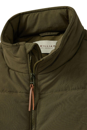 A close-up of the RM Williams Padstow Vest in olive green, showcasing its high collar and zip closure accentuated by a brown leather pull tab. The slightly visible interior label reveals the brand name. This vest is made from recycled nylon with a quilted design and is filled with PrimaLoft, ensuring warmth and insulation.