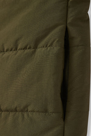 Close-up view of the RM Williams Padstow Vest made from green quilted recycled nylon, showcasing visible stitching that forms rectangular sections. A partially visible black zipper is located on the right side of the image, with the texture suggesting it's part of a high-quality puffer vest featuring PrimaLoft filling.