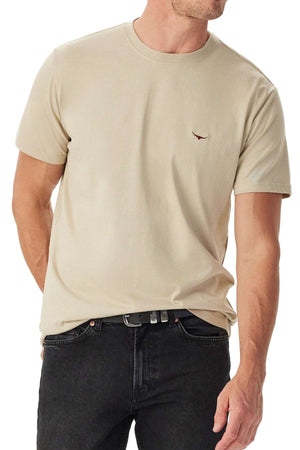 A man wears the beige RM Williams Parson T-Shirt, featuring a small embroidered chest logo. He pairs it with black jeans and a black belt, standing slightly side-on with one arm relaxed.