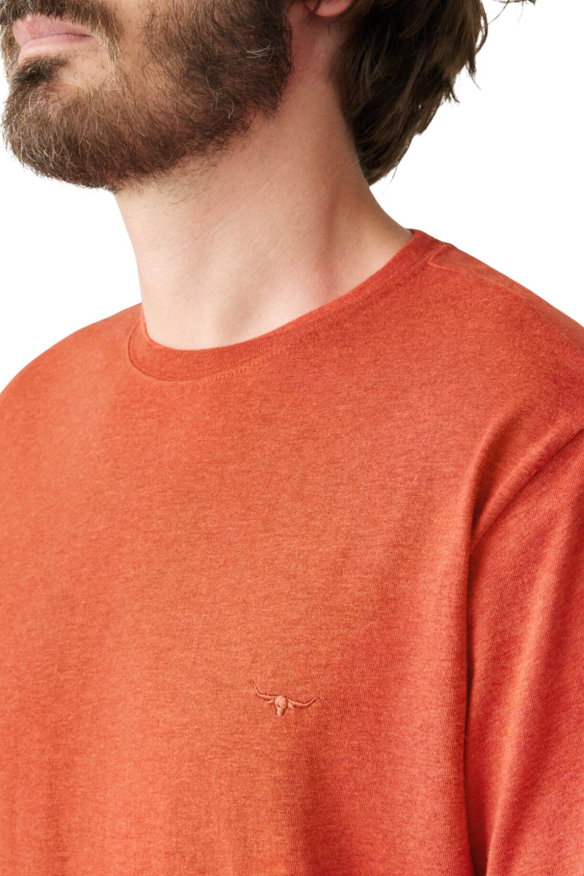 A person with a beard is wearing the RM Williams Parson T-Shirt, an orange, organic cotton crew neck top featuring a small embroidered horns logo on the chest. The image is cropped to show the upper body and part of their face, highlighting its casual style.