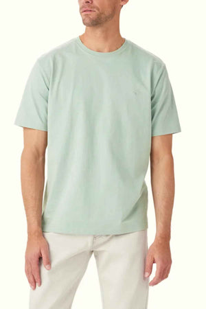 A man wearing an RM Williams Parson T-Shirt in light green with a crew neck, made of organic cotton, along with beige pants, stands against a plain background.