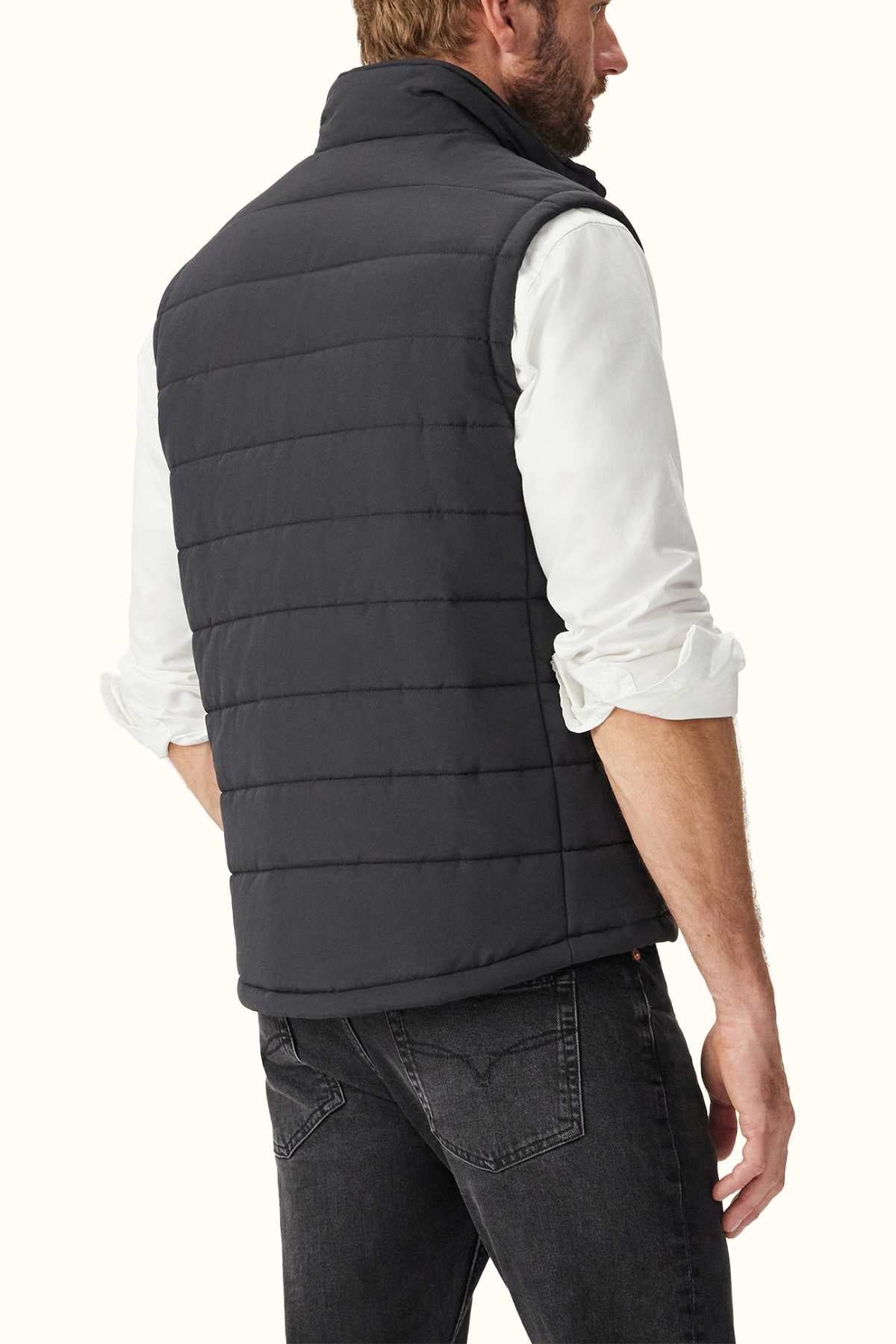 A man with short hair stands facing away from the camera, wearing a white long-sleeve shirt and the RM Williams Patterson Creek Vest made of recycled polyester, paired with dark jeans. The lightweight padding of the dark vest adds style to the plain, light-colored background.