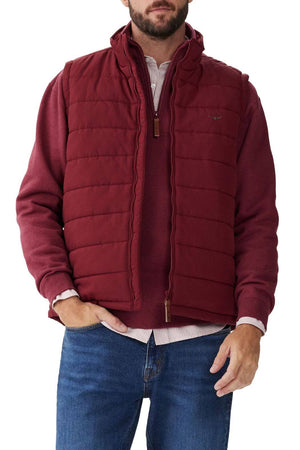 A person is wearing an RM Williams Patterson Creek Vest in maroon over a matching maroon long-sleeve shirt. They are also wearing blue jeans and standing with one hand in their pocket. The water-resistant, quilted puffer vest features a stand-up collar and a zip closure. The background is plain white.