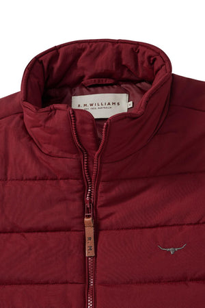 A close-up of a maroon RM Williams Patterson Creek Vest showcases its water-resistant material and high collar. The vest is designed with a zipper that has a brown leather pull tab, along with a small, embroidered logo of a bull's head on the chest. Inside the collar, a label displaying the brand name R.M. Williams is visible.