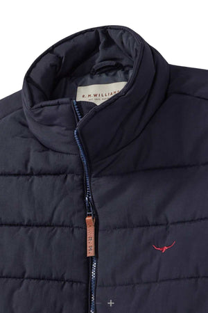 A close-up view of the RM Williams Patterson Creek Vest, a black quilted vest with a high collar crafted from recycled materials. This lightweight vest features a brown zip puller with embossed text and a small embroidered red longhorn logo on the chest. The inside label reads "R.M. Williams.