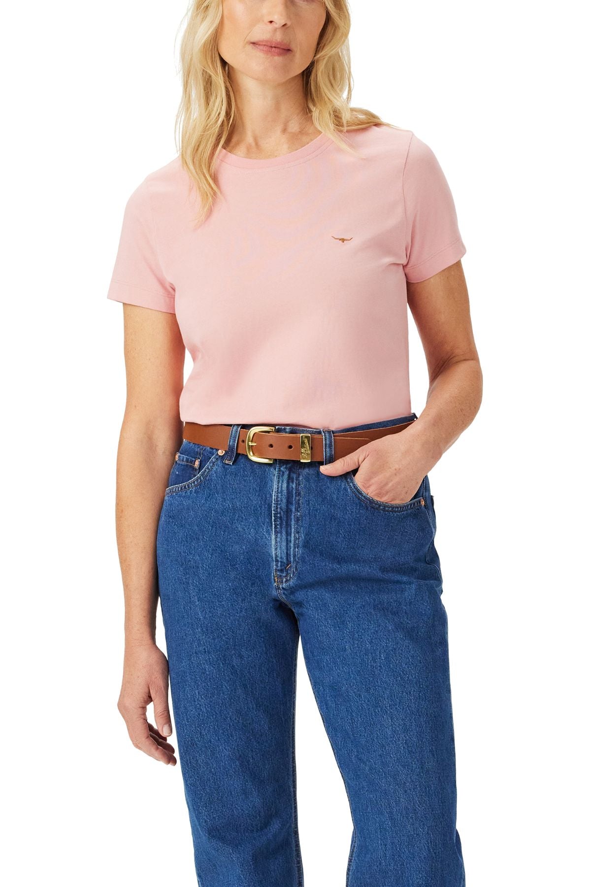 A woman wears the RM Williams Piccadilly Tee in light pink, tucked into high-waisted blue jeans with a brown belt, standing with one hand in her pocket.