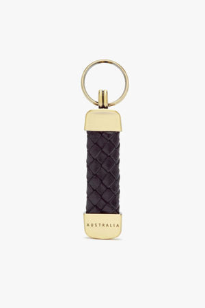 The RM Williams Plaited Key Ring features a gold ring with a woven black Kangaroo leather handle, accented by brass-toned fittings and engraved "AUSTRALIA" metal ends. The design is simple and elegant against a plain white background.