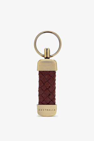 The RM Williams Plaited Key Ring features a brass keyring with a brown braided kangaroo leather body, metallic end caps, and "AUSTRALIA" engraved on the bottom, combining minimalist design with premium brass-toned fittings.