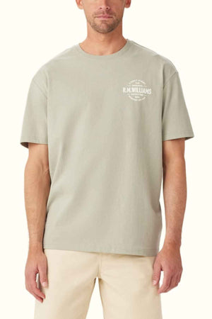 A man wearing a light gray RM Williams Prospect T-Shirt, featuring a classic fit and a subtle graphic print logo on the chest. Made from organic cotton, this shirt is paired with beige pants. The plain background maintains the focus on the simplicity and style of the clothing.