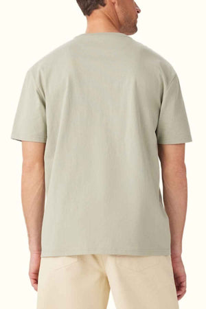 A man is standing with his back to the camera, highlighting the RM Williams Prospect T-Shirt in light gray organic cotton with a classic fit, paired with beige pants. The plain off-white background subtly allows the fabric's texture to stand out.