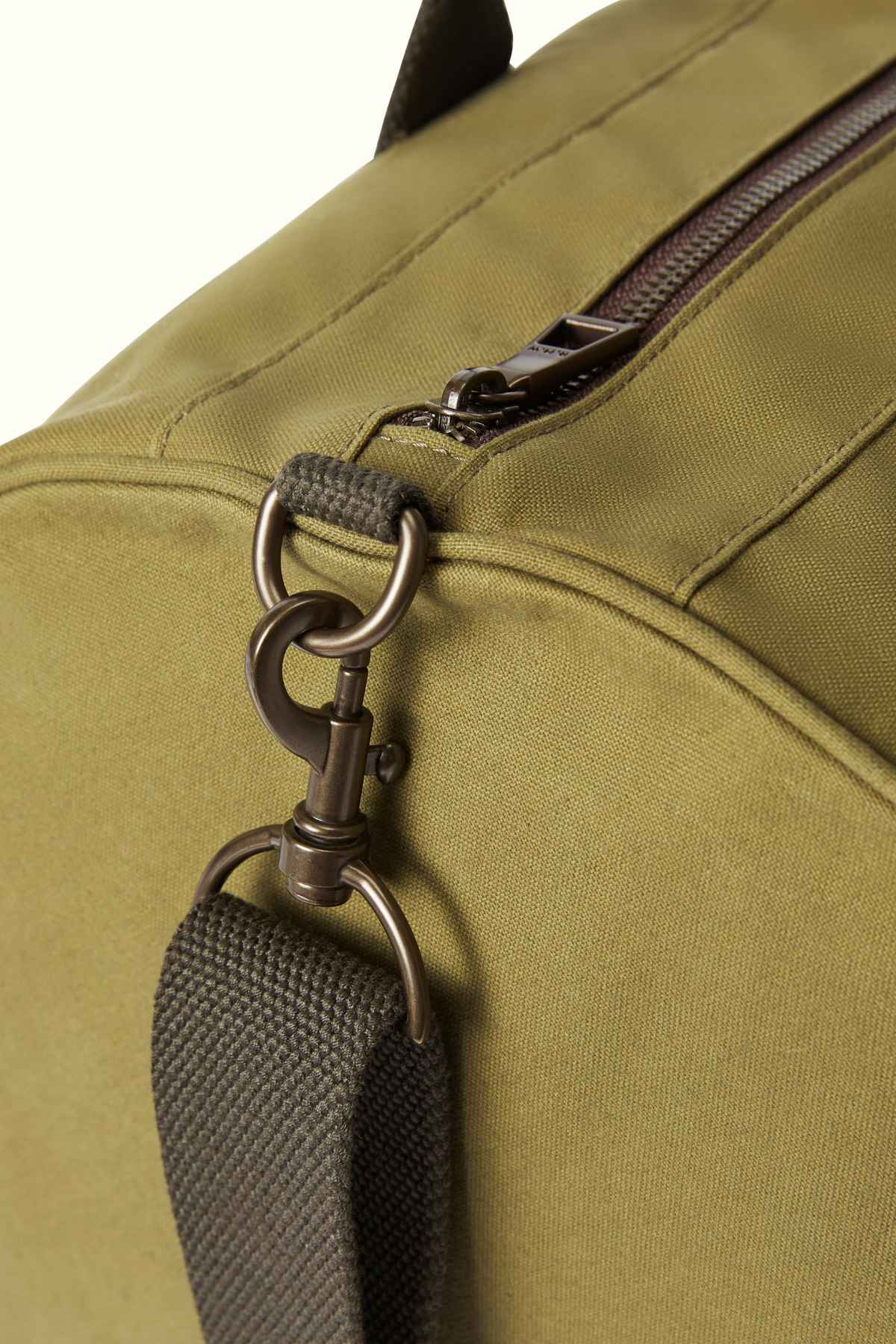 This close-up showcases the RM Williams RM Ditty Bag in olive-green, crafted from waxed cotton canvas. It highlights a metal clasp and hook attaching a fabric strap, with a visible top zipper that underscores its sturdy construction and functional design. The bag is made by the esteemed manufacturer Halley Stevensons.