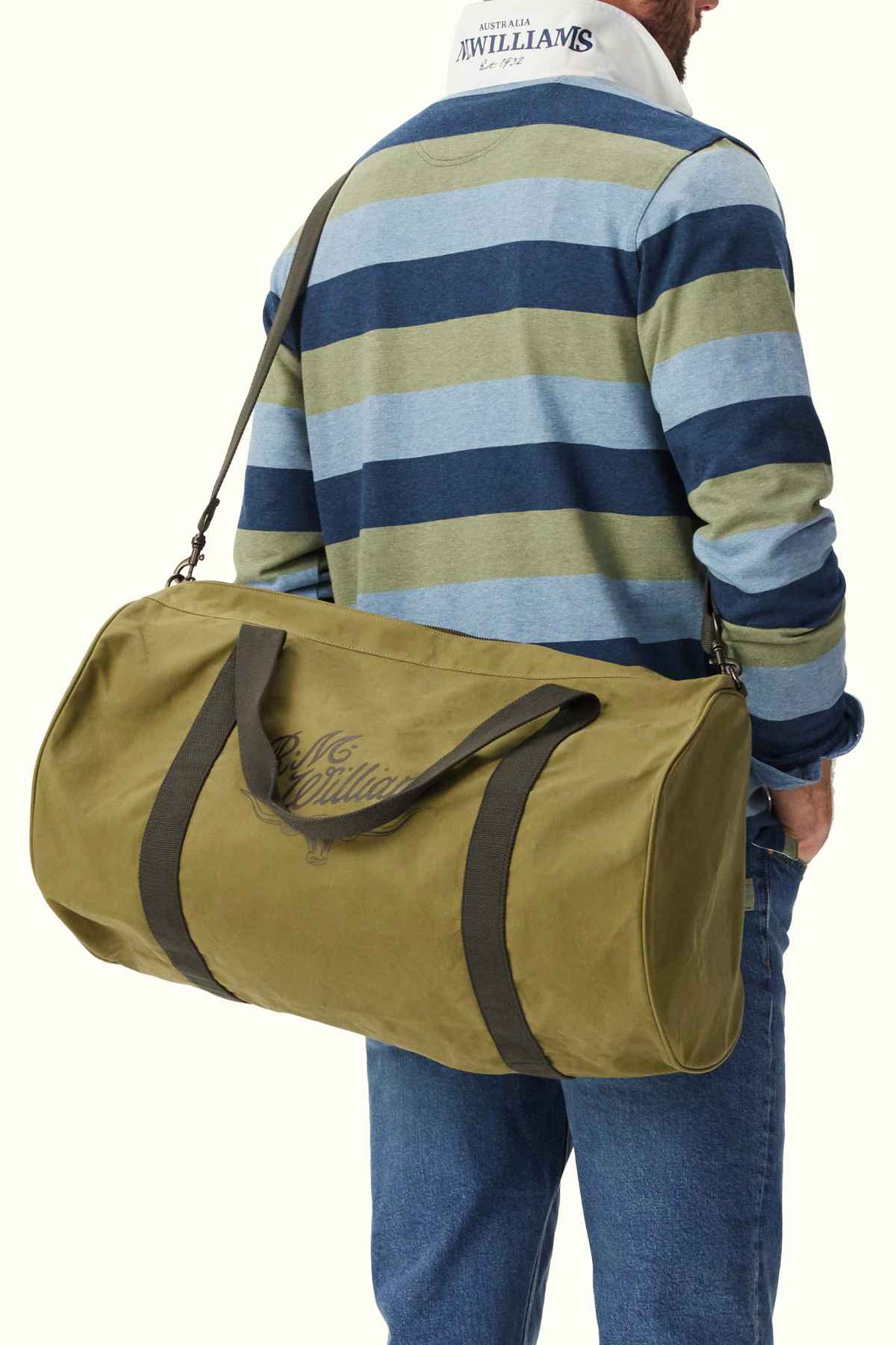A man wearing a striped rugby shirt and blue jeans is carrying an RM Williams RM Ditty Bag—an olive green duffel crafted from durable waxed cotton canvas—over his shoulder. The shirt proudly displays the R.M. Williams logo on the back collar, making him and his bag perfect travel companions.
