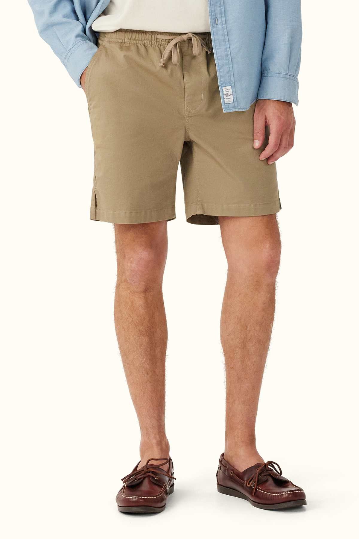 A person wearing RM Williams R.M.W Rugby Shorts in khaki, an elasticated waist, a light blue button-up shirt slightly tucked in, and brown leather boat shoes with tassels against a neutral background.