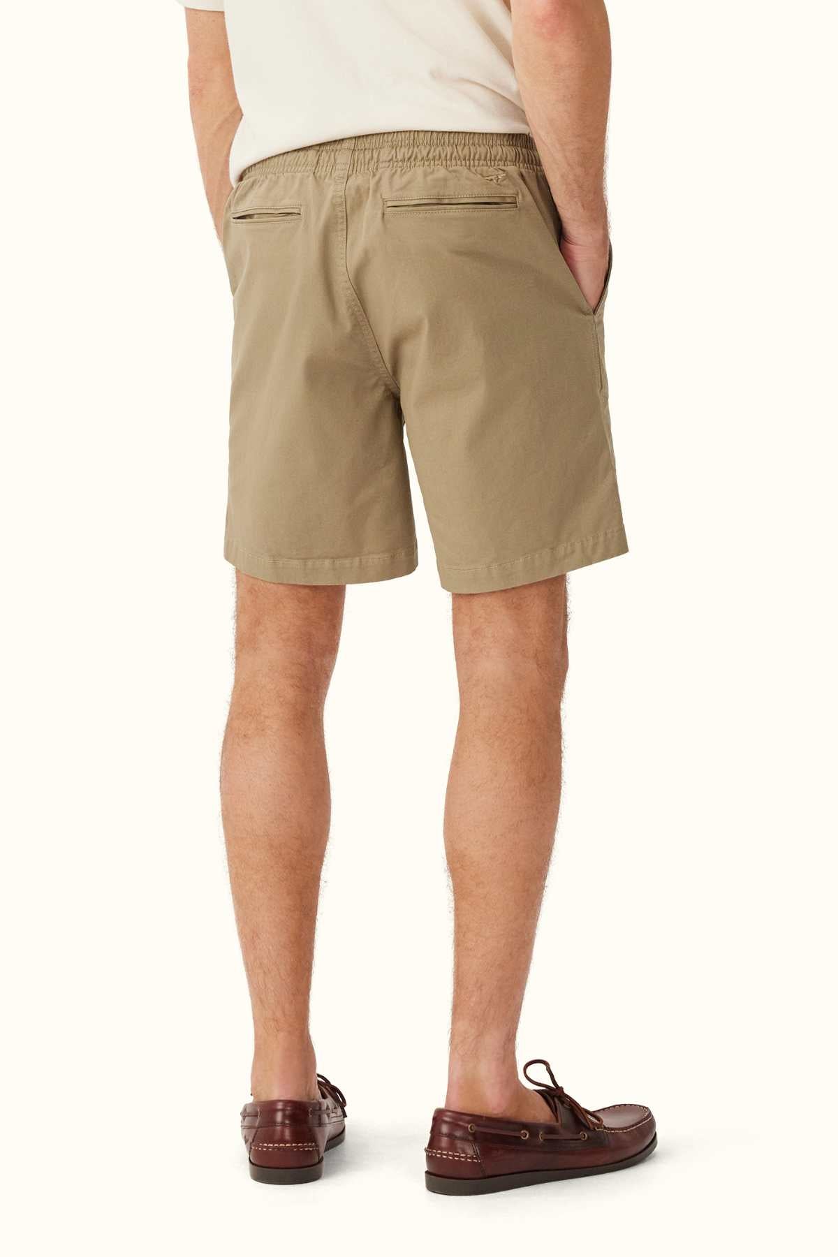 A person wearing a beige T-shirt and RM Williams R.M.W Rugby Short in khaki stands with their back to the camera. These shorts, crafted from washed cotton twill, are designed with an elasticated waist for added comfort. With hands in pockets and brown boat shoes completing the look, they stand against a light background.