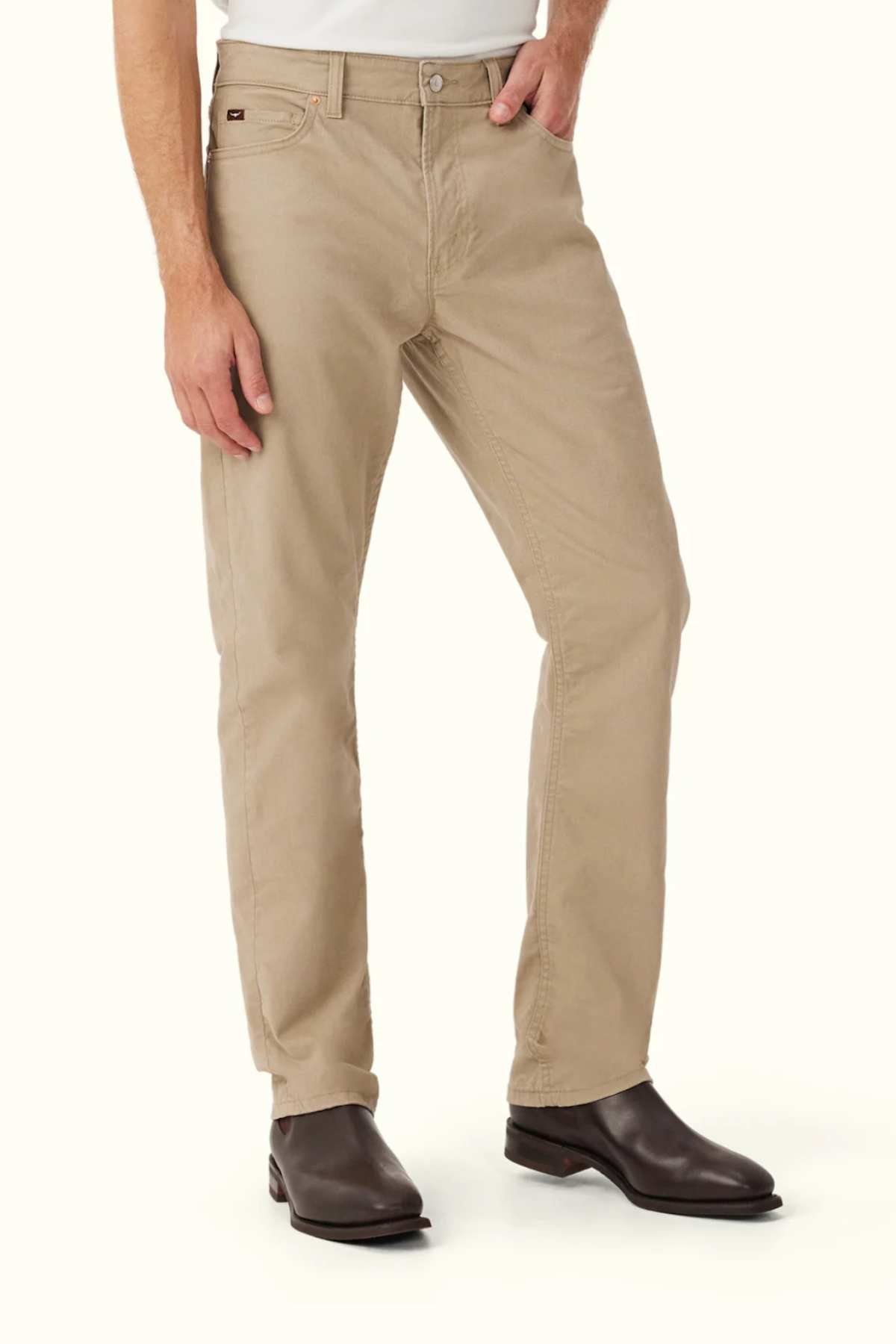 A person wearing RM Williams Ramco 5 Pocket Jeans in beige stretch cotton twill, a white shirt, and dark brown leather boots stands with one hand in their pocket against a plain light background.