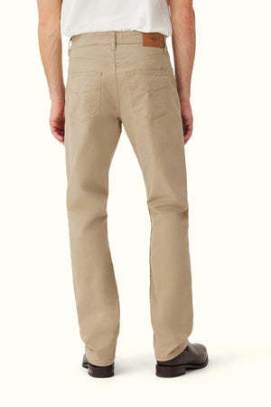 Someone stands with their back to the camera against a plain background, wearing beige RM Williams Ramco 5 Pocket Jeans made from Australian cotton, paired with a white shirt and dark brown shoes.