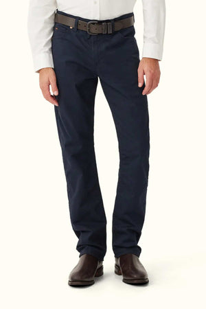 A man stands against a plain light background, shown from shoulders to feet, wearing a white long-sleeve shirt and RM Williams Ramco 5 Pocket Jeans in dark navy, paired with a brown belt and brown dress shoes.